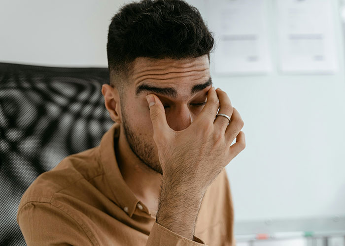 Man making family uncomfortable, sitting in a chair, rubbing eyes with a frustrated expression in a home setting.