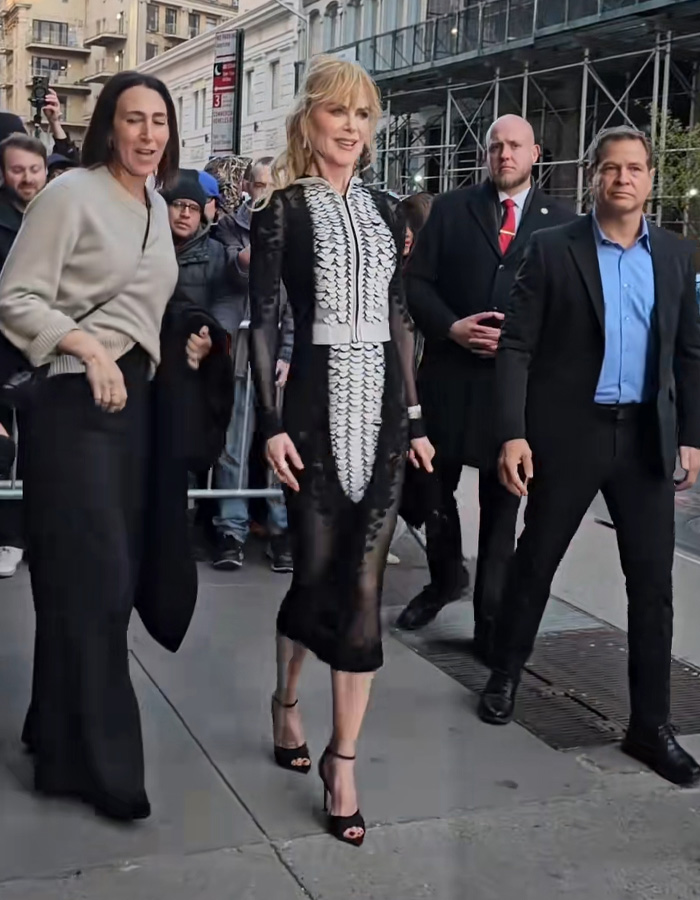 Nicole Kidman wearing a sheer black and white dress at a premiere, walking with people around her on city sidewalk.