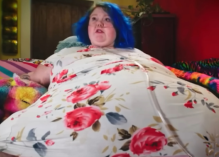 My 600-Lb Life star Dolly Martinez with blue hair and floral dress lying on a colorful bed in a home setting.