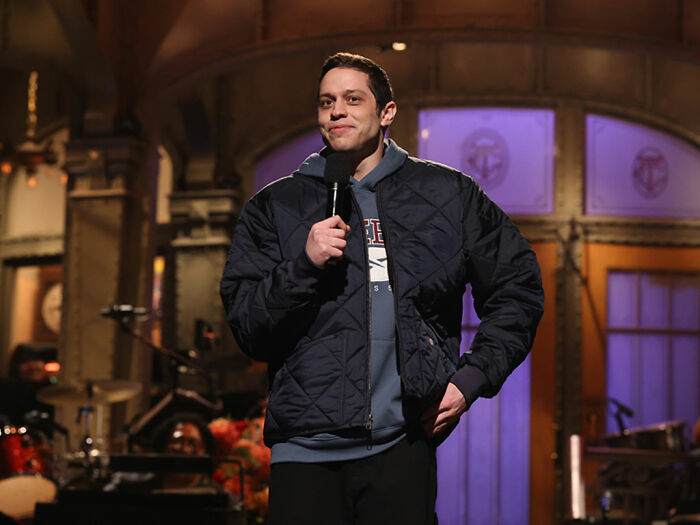Pete Davidson performing stand-up comedy on stage, holding a microphone, sparking debate about adult content and fatherhood.
