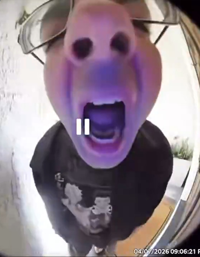 Intruso visto em vídeo gritando para a câmera da campainha, close-up com óculos e roupas escuras na entrada da residência.