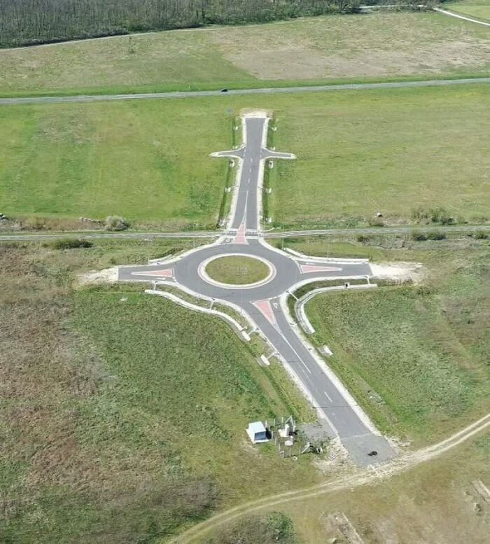 Aerial view of a weird image: a cross-shaped road leading to a roundabout in the middle of a green field.