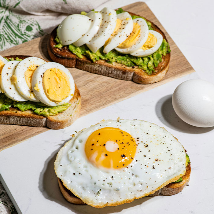 Torradas de abacate cobertas com ovos fatiados e fritos, acompanhando um ovo cru. Comer ovos para tratamento de Alzheimer.