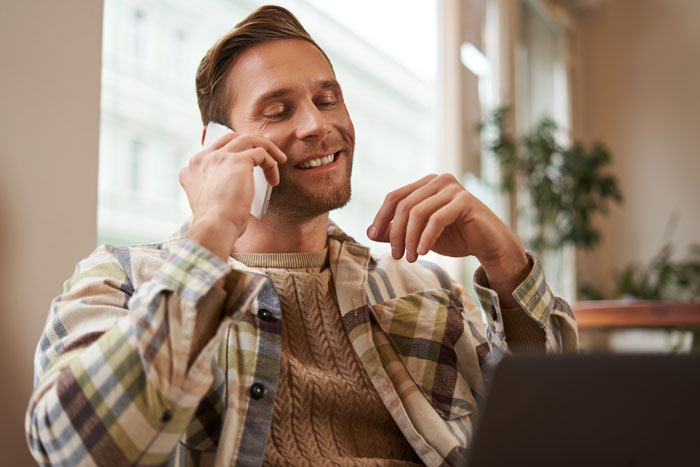 A man in a plaid shirt and knit sweater smiling, talking on a smartphone, perhaps having a dumb dating moment.