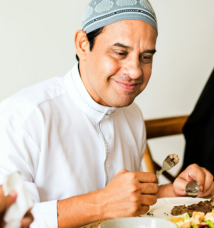 Um homem muçulmano vestindo kufi e thobe branco sorri enquanto come, sem saber que contém gordura de bacon.