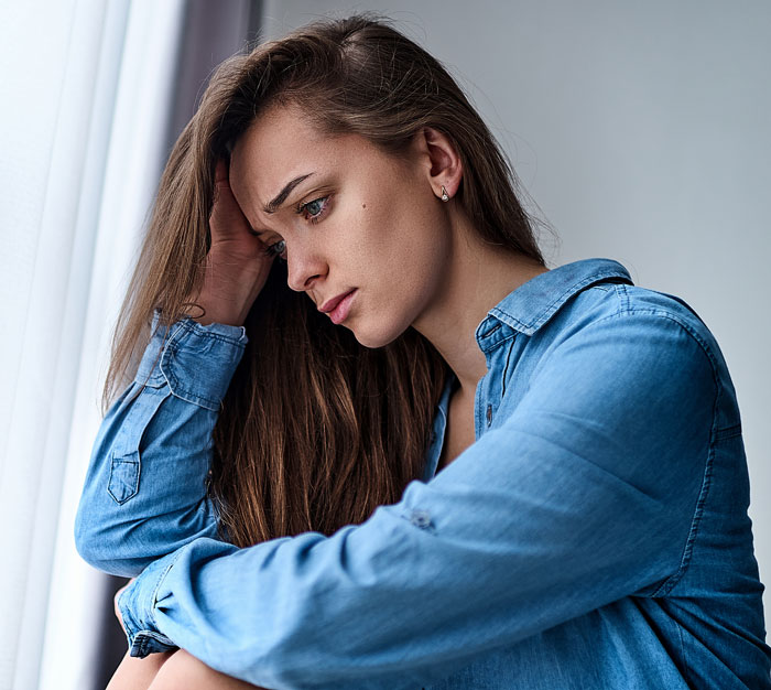Pensive woman in a blue shirt sitting by a window, reflecting on things women are tired of explaining to men.
