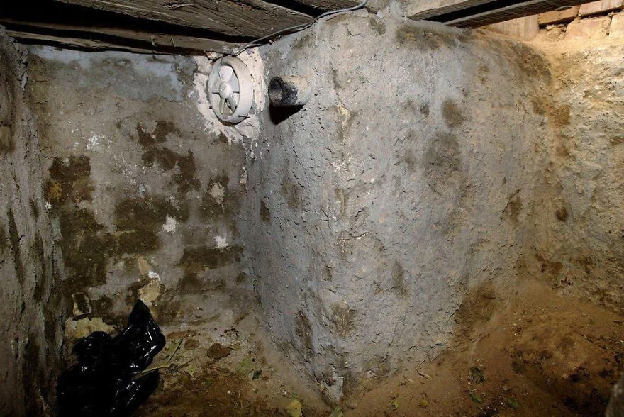 A dark, damp basement with concrete walls, a small vent, and dirt floors. This image suggests a place with tough security.