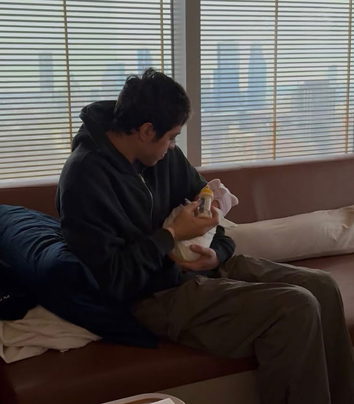 Man feeding baby with bottle on couch near large window, representing Pete Davidson parenting with daughter topic.