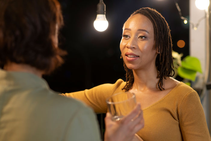 A Black woman with dreadlocks in a yellow top holding a glass, talking to someone at a night gathering about Black people.