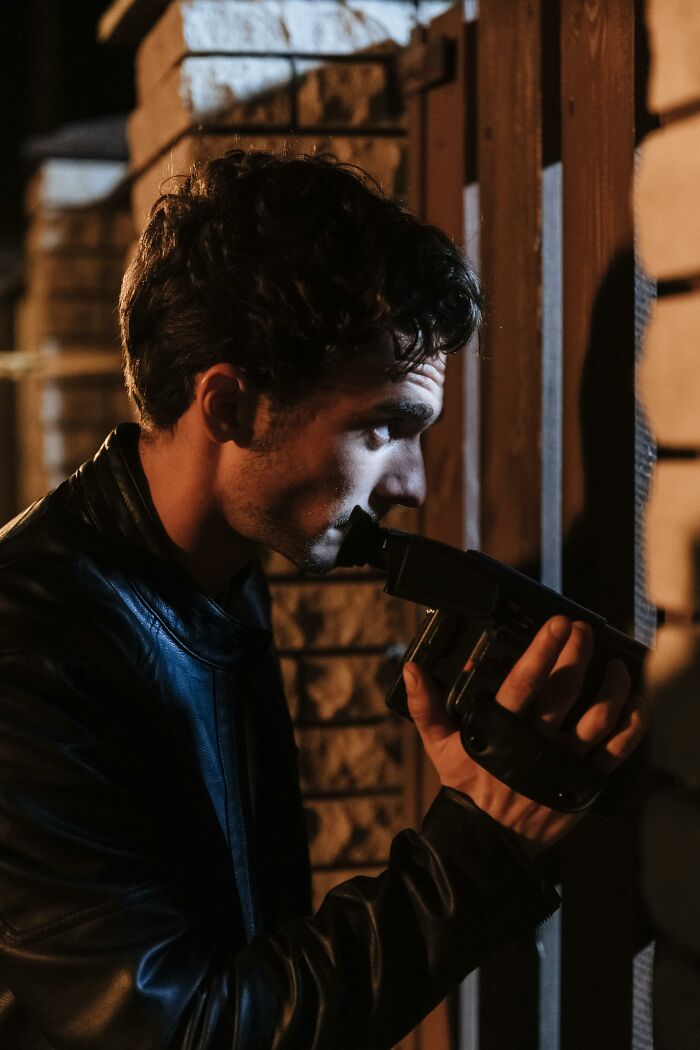 A man in a leather jacket holds a camcorder, peeking around a corner, hinting at workplace incidents.