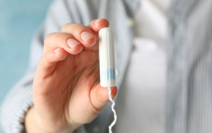 A hand holds up a white tampon with a blue band, illustrating a gross office story.