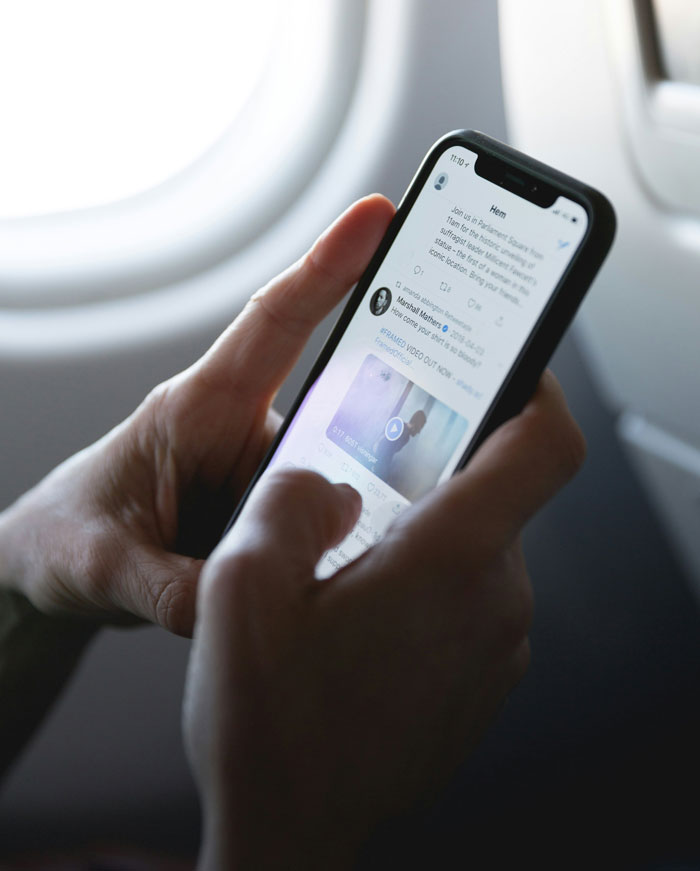 Hands hold a smartphone scrolling through social media, a plane window in the background. Reflecting on dating someone dumb.