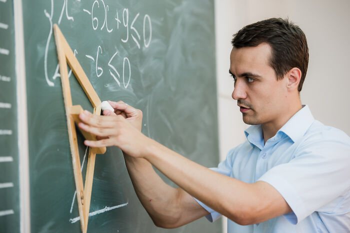 A man in a light blue shirt uses a large wooden compass to draw on a blackboard with chalk, sharing wild rumors.