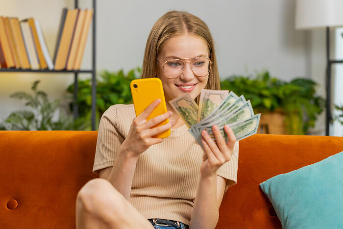 Young woman on orange couch holding cash and yellow phone, symbolizing inspiring comebacks and thriving success.