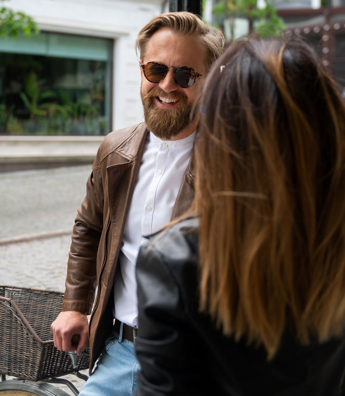 Bearded man wearing sunglasses and a brown leather jacket smiling while talking to a woman outdoors, petty karma concept.