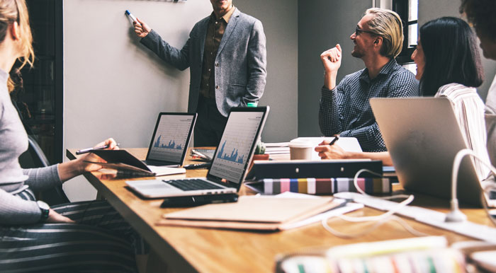 A group of professionals in a meeting, with laptops showing graphs. Presenter points at a board, discussing Toxic Bosses strategies.