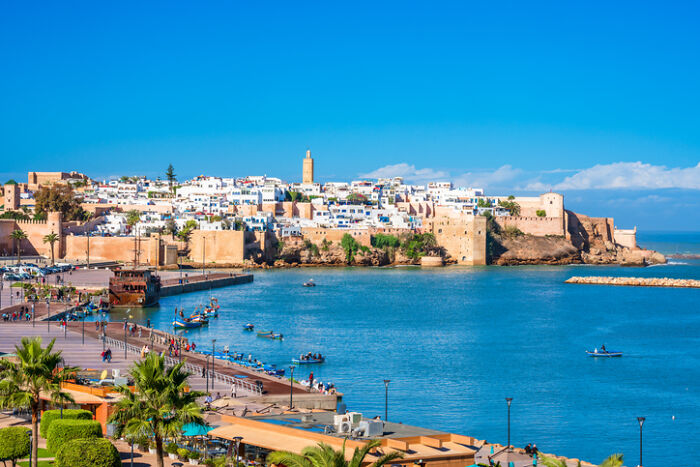 Coastal city view with white buildings, ancient walls, and boats on blue water. Represents countries with worst work-life balance.
