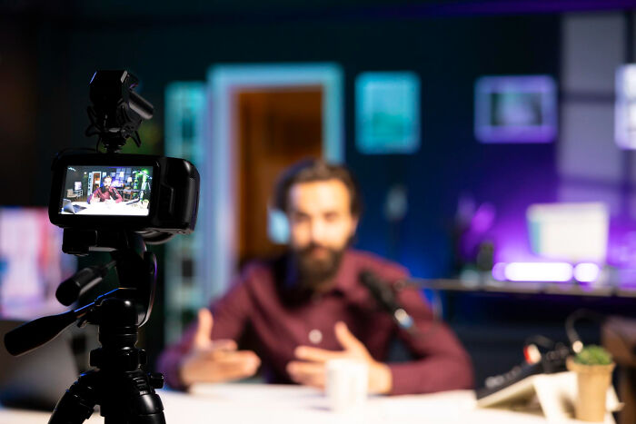 A man with a beard records a video, seen on the camera screen, sharing stories about celebrity relatives.