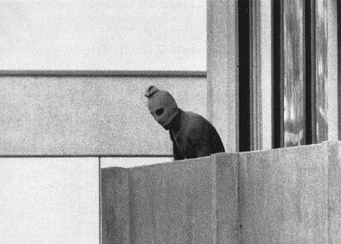 Black and white photo from 1960 to 2010 showing a person in a mask on a building balcony during a notable event.