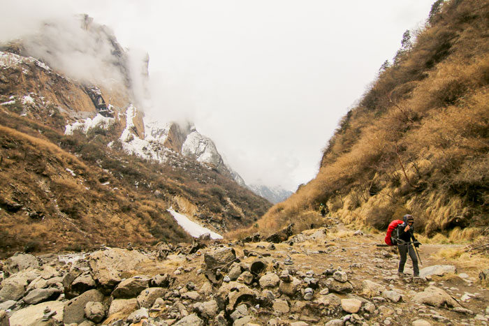 Hiker trekking through rocky mountainous terrain with dry trees and snow patches under cloudy sky.