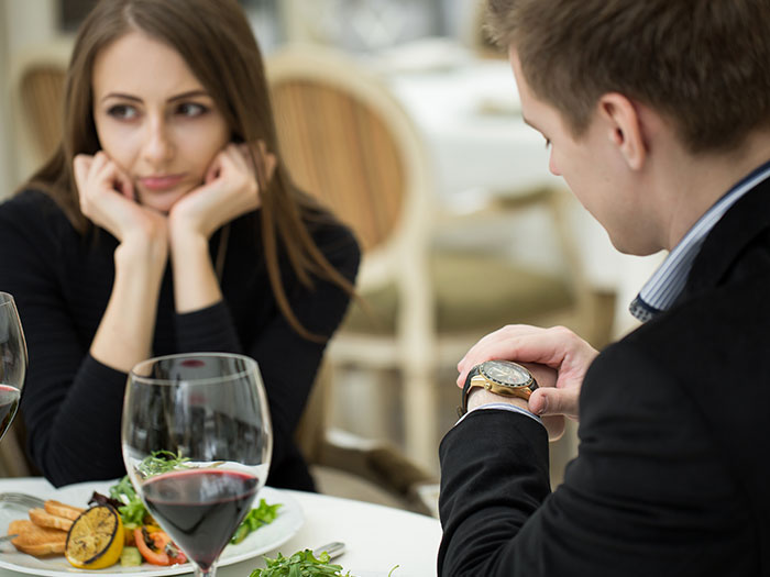 A man checking his watch while a woman looks bored during a first date, emphasizing a horrible first date experience.