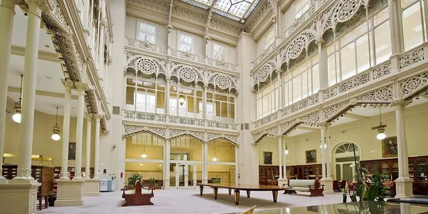 A grand, ornate interior hall with multiple levels, intricate white railings, columns, and a glass ceiling. Toughest security.