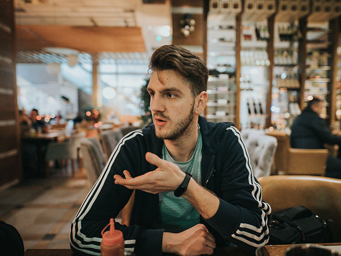 A man in a casual tracksuit jacket at a restaurant, gesturing as if telling a story about horrible first dates.