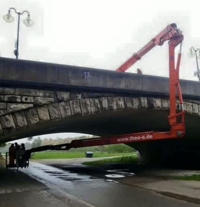 A weird image of an orange boom lift struggling to reach over and under a stone bridge, making people go hmmm.