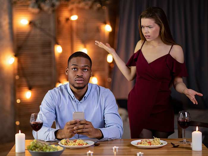 A man on his phone during a horrible first date, while a frustrated woman gestures behind him at a dimly lit restaurant.