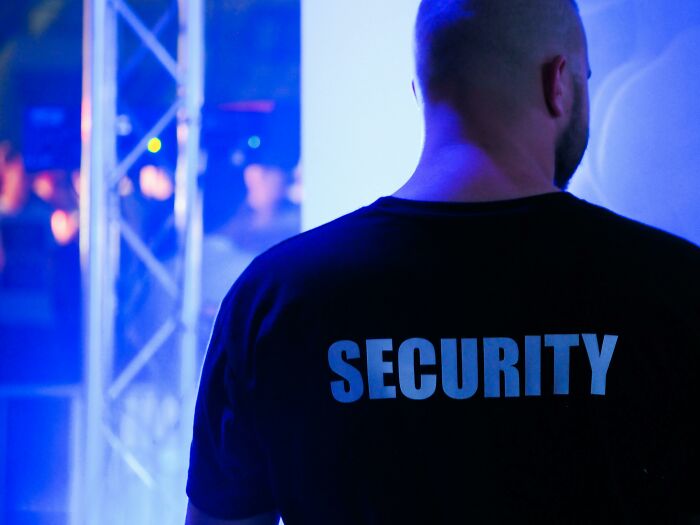 A security guard's back, facing blue light with "SECURITY" on his shirt. Workplace incidents require such protection.