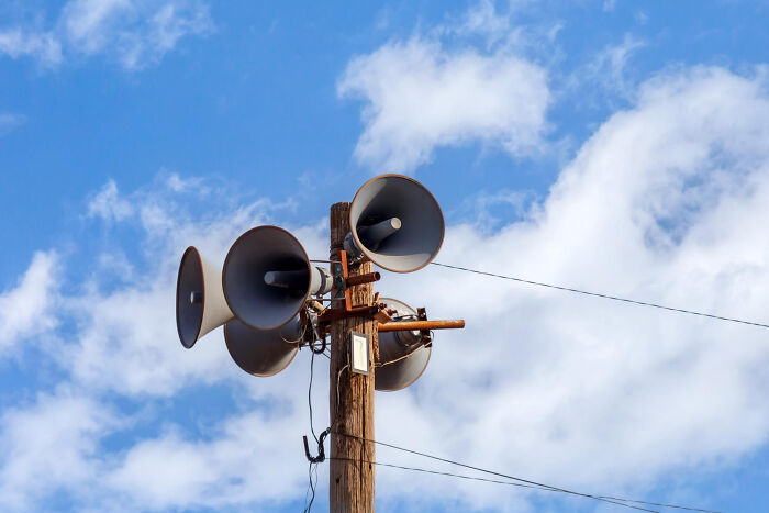 Four large, grey loudspeakers mounted on a wooden utility pole against a cloudy blue sky. Disturbing sounds.