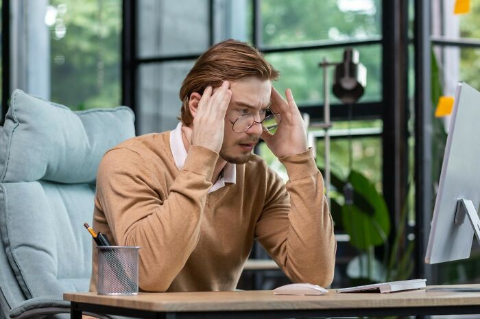 A man with glasses and a brown sweater, hands on his temples, looking stressed at a computer, hearing wild rumors.