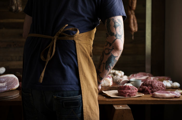 Rear view of a person with a tattooed arm and brown apron, preparing raw meat on a rustic table, potentially for shared workspace.