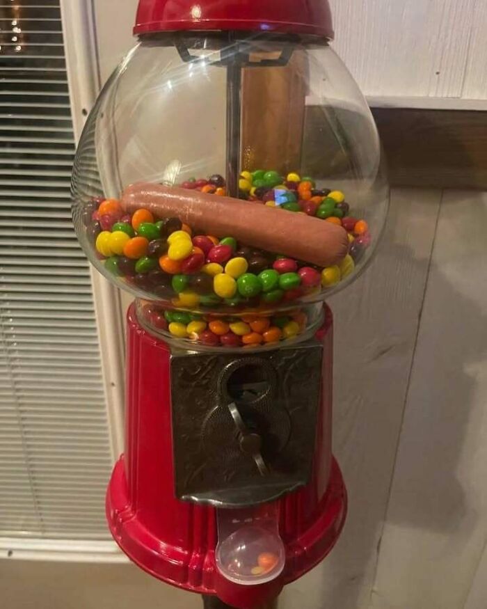 A confusing and unhinged image of a red gumball machine filled with colorful candy and a single hotdog inside.