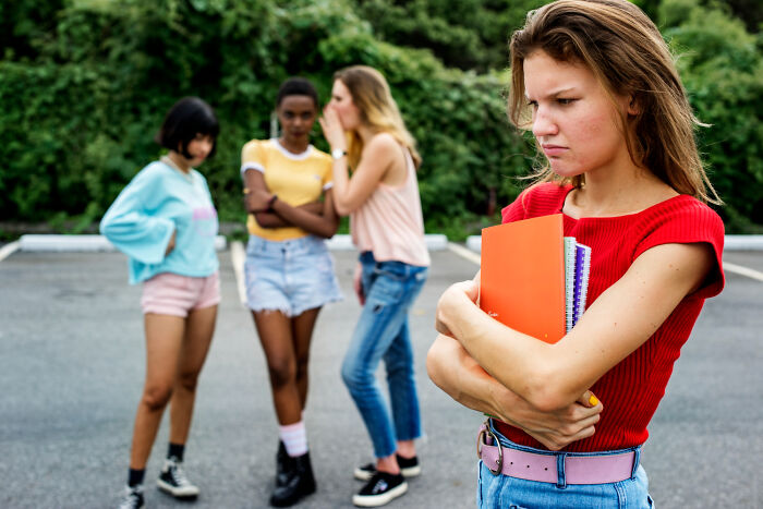Teen girl holding notebooks and looking upset while three other girls whisper and gossip behind her, depicting bullying experience.