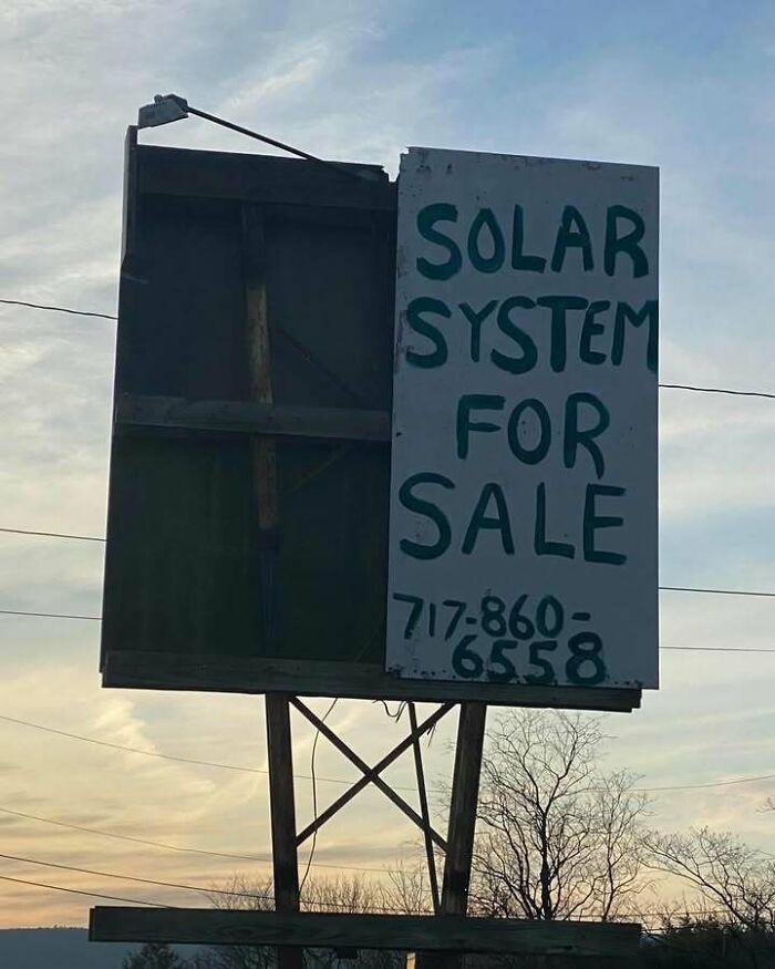 Handwritten sign on billboard reading solar system for sale with phone number under cloudy sky, a confusing and unhinged image.