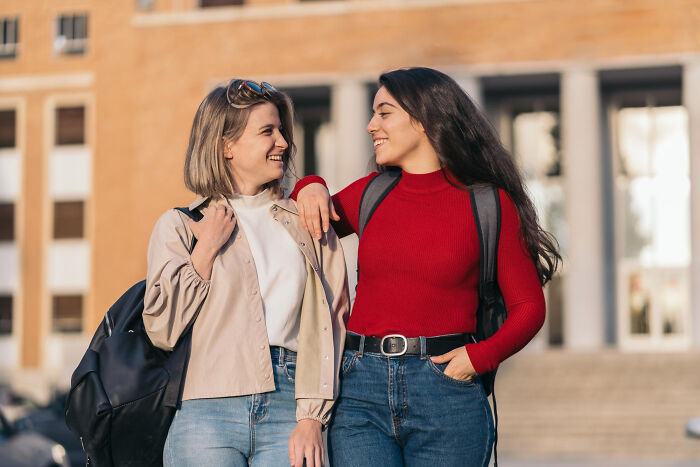 Two happy female students with backpacks outside, smiling at each other. They could be sharing wild rumors.