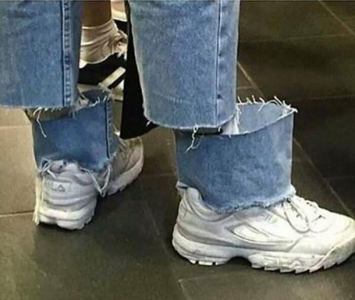 Frayed jeans cuffs unusually tucked inside white sneakers, creating a confusing and unhinged fashion look on tiled floor.