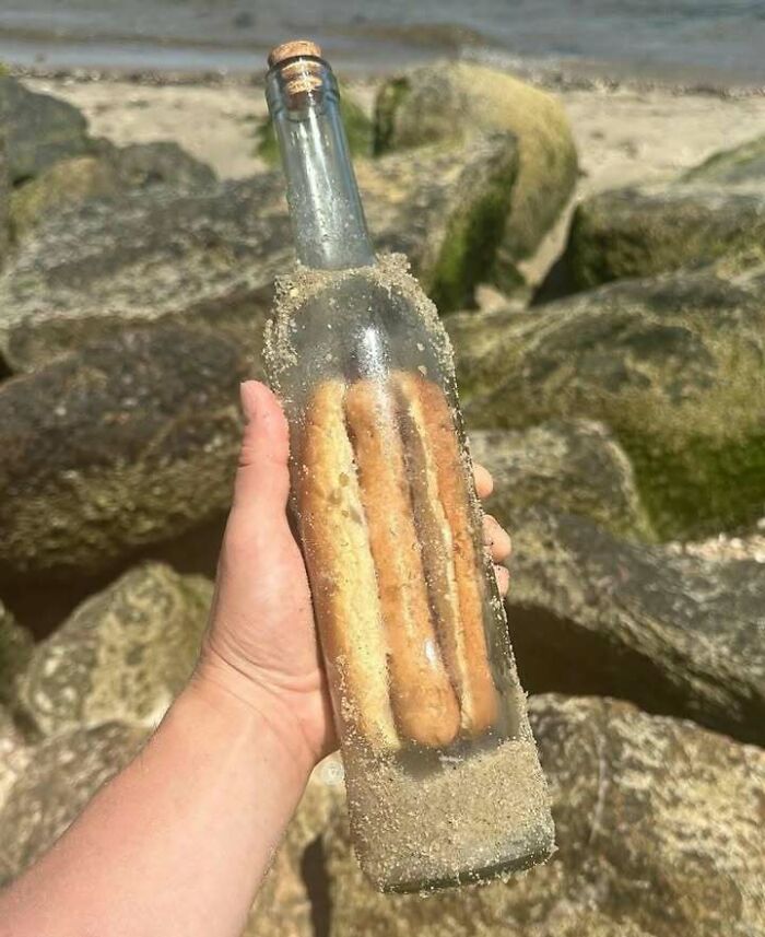 Hand holding a bottle filled with three breadsticks covered in sand, a confusing and unhinged image by the shore.