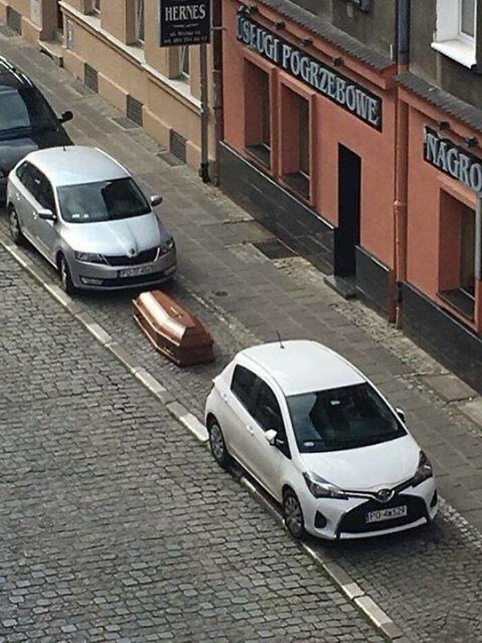 Coffin placed on sidewalk between two parked cars on a cobblestone street in a confusing and unhinged scene.