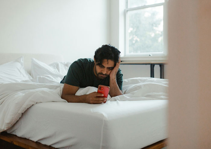 Man looking stressed and lying on bed using phone, illustrating oversleeping turning into a parenting nightmare moment.