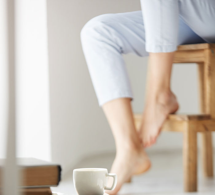 A person's lower legs and feet, one on a wooden stool, with a coffee cup and books below. Relates to office stories.