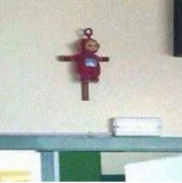 Blurry Teletubby toy hung on a cross-shaped wooden plaque on a plain wall in a confusing and unhinged image.