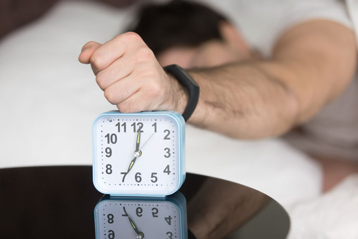 Person in bed turning off alarm clock, illustrating the stress of oversleeping and parenting struggles.