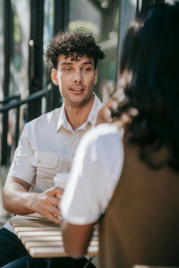 A man with curly hair and a light shirt, holding a coffee, talking to a woman across a table. A first date.