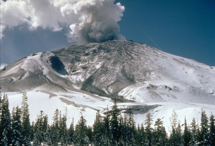 Snow-covered volcano erupting with smoke plume rising above trees, representing major events in the year you were born.