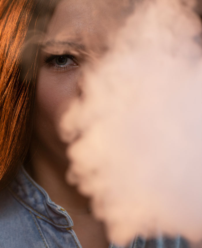 Close-up of a woman's face, half-obscured by smoke. Her blue eye peers through, highlighting gross office stories.