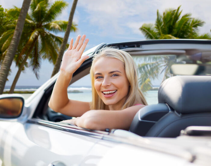 Smiling woman waving from convertible car near palm trees, symbolizing inspiring comebacks and thriving success.
