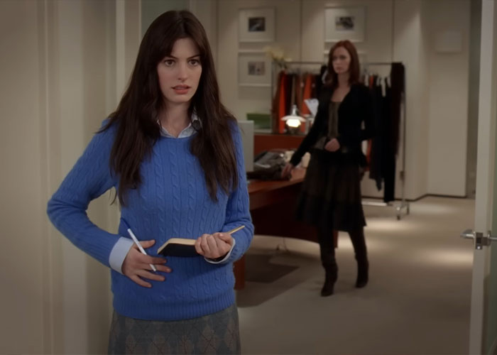 Woman in blue sweater holding a notebook with another woman in the background portraying Vogue office dynamics and assistants.