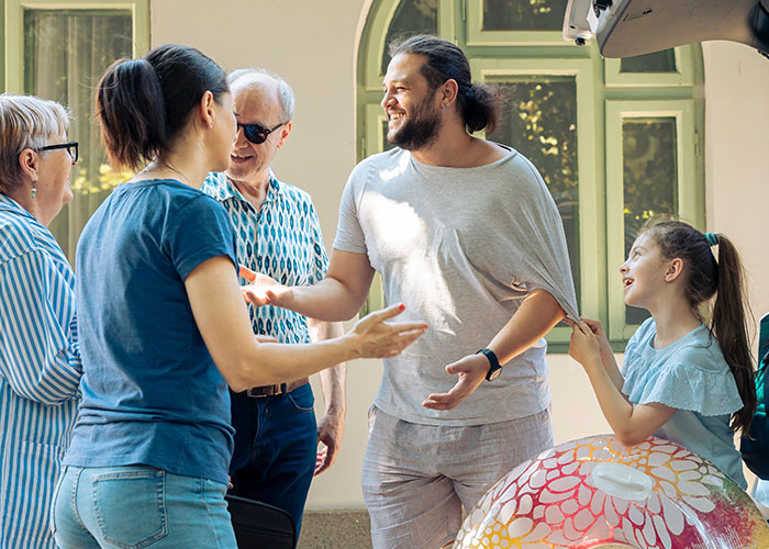 Man making family uncomfortable at home while they interact near an open car trunk during a sunny day.
